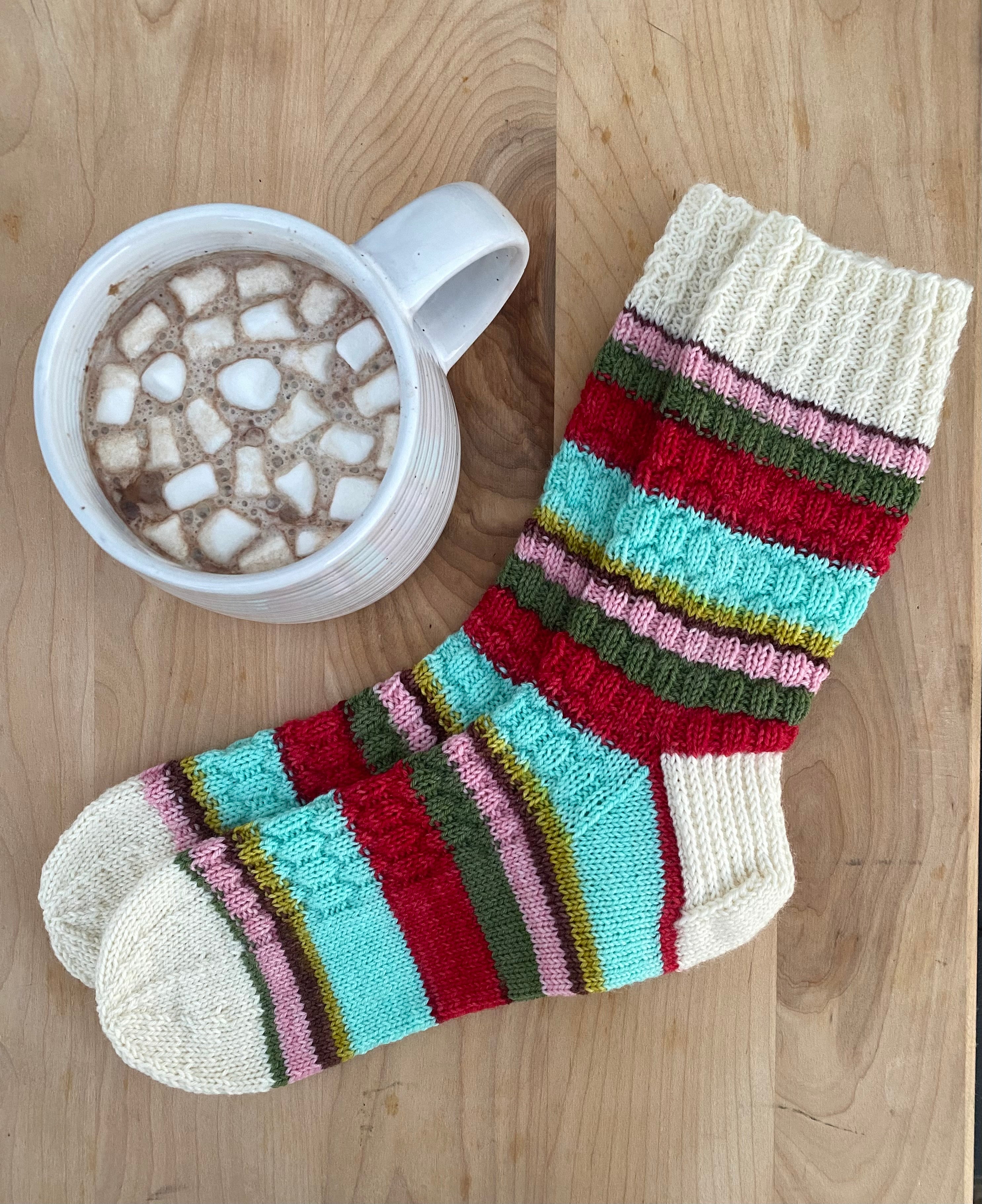 A pair of colorful knitted socks next to a cup of hot cocoa on a wooden surface, representing the 'Nutcracker' themed product.